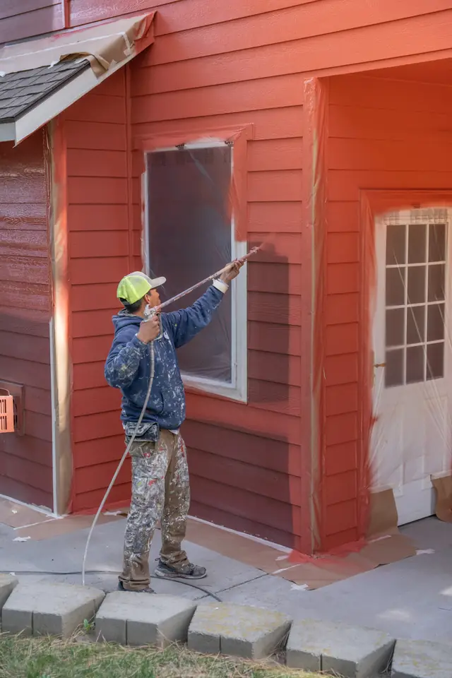 A painter in a mask and cap spraying red siding, with the windows masked in paper