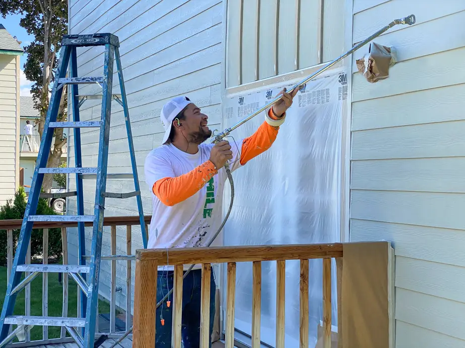 A painter in a Smile-A-Mile tee spraying pale siding from a ladder, windows masked in plastic